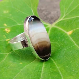 May include: A silver ring with a large, oval stone. The stone has bands of white, tan, and dark brown. The ring is set on a bright green leaf.