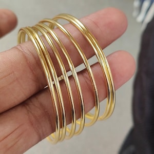 Op de afbeelding: Een set van zes goudkleurige armbanden. De armbanden zijn dun, rond en lijken van metaal te zijn. De armbanden zijn op elkaar gestapeld en het licht reflecteert op hun gladde oppervlakken. De sieraden worden gepresenteerd tegen een neutrale achtergrond.