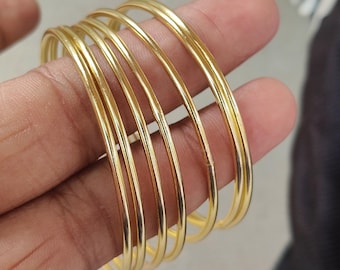 以下が含まれることがあります： 6つの金色のバングルセット。バングルは細く丸く、金属製に見えます。バングルは積み重ねられており、光が滑らかな表面に反射しています。ジュエリーはニュートラルな背景に表示されています。