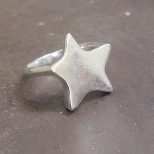 Könnte beinhalten: Ein silberner Ring mit einem fünfzackigen Stern-Design. Der Stern ist der Mittelpunkt, mit einer glatten, leicht strukturierten Oberfläche. Der Ring ist schlicht und poliert, was eine minimalistische Ästhetik erzeugt.