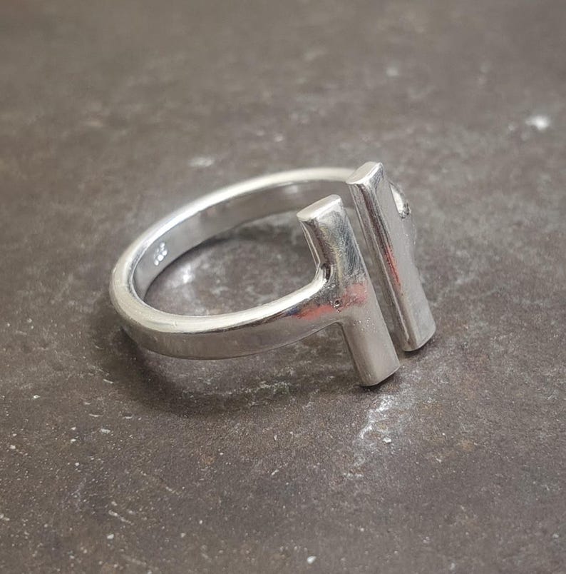 May include: A silver ring with a modern design. The ring features a band and two vertical rectangular bars that form a T-shape. The ring is made of a shiny, reflective metal and is set against a dark, textured surface.