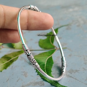 May include: A silver bangle bracelet with detailed, textured sections. The bracelet has a curved, open design with decorative bands near the ends. The background includes green leaves and a blue-grey surface. The text "silverringartisanco" is visible.