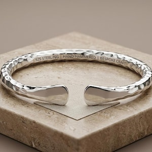 Può includere: Un bracciale a fascia in argento con una finitura martellata a mano. L'interno del cinturino presenta il testo inciso 925 STERLING | HAND-HAMMERED | BESPOKE CRAFT. Il bracciale ha un design aperto con due estremità lisce e arrotondate.