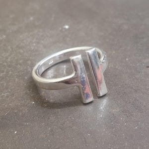 May include: A silver ring with a modern design. The ring features two vertical bars connected to the band, creating an open, geometric shape. The ring is made of a shiny, metallic material.
