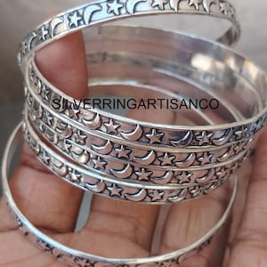 Peut inclure: Plusieurs bracelets argentés ornés d'un motif céleste. Chaque bracelet présente un motif répétitif de croissants de lune et d'étoiles. Le texte "SILVERRINGARTISANCO" est visible sur l'image.