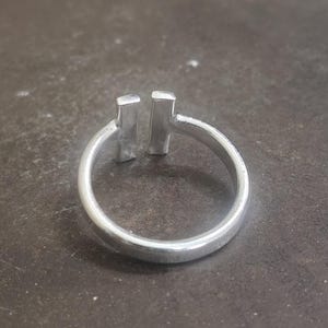 May include: A silver ring with a minimalist design. The ring features a smooth, rounded band and two vertical rectangular bars set close together at the top. The ring is made of a shiny, metallic material.
