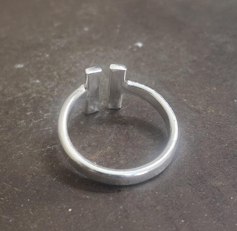 May include: A silver ring with a minimalist design. The ring features a smooth, rounded band and two vertical rectangular bars set close together at the top. The ring is made of a shiny, metallic material.