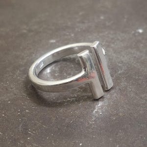 May include: A silver ring with a modern design. The ring features a band and two vertical rectangular bars, creating a unique T-shape. The ring is made of a shiny, metallic material and is set against a dark, textured surface.