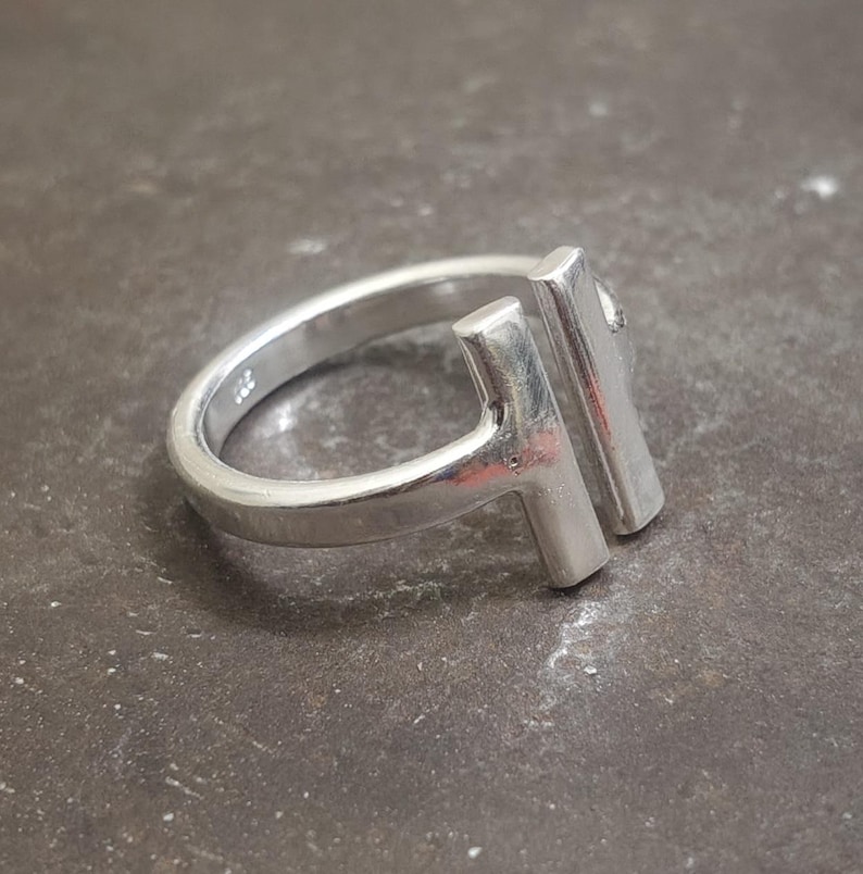 May include: A silver ring with a modern design. The ring features a band and two vertical rectangular bars, creating a unique T-shape. The ring is made of a shiny, metallic material and is set against a dark, textured surface.