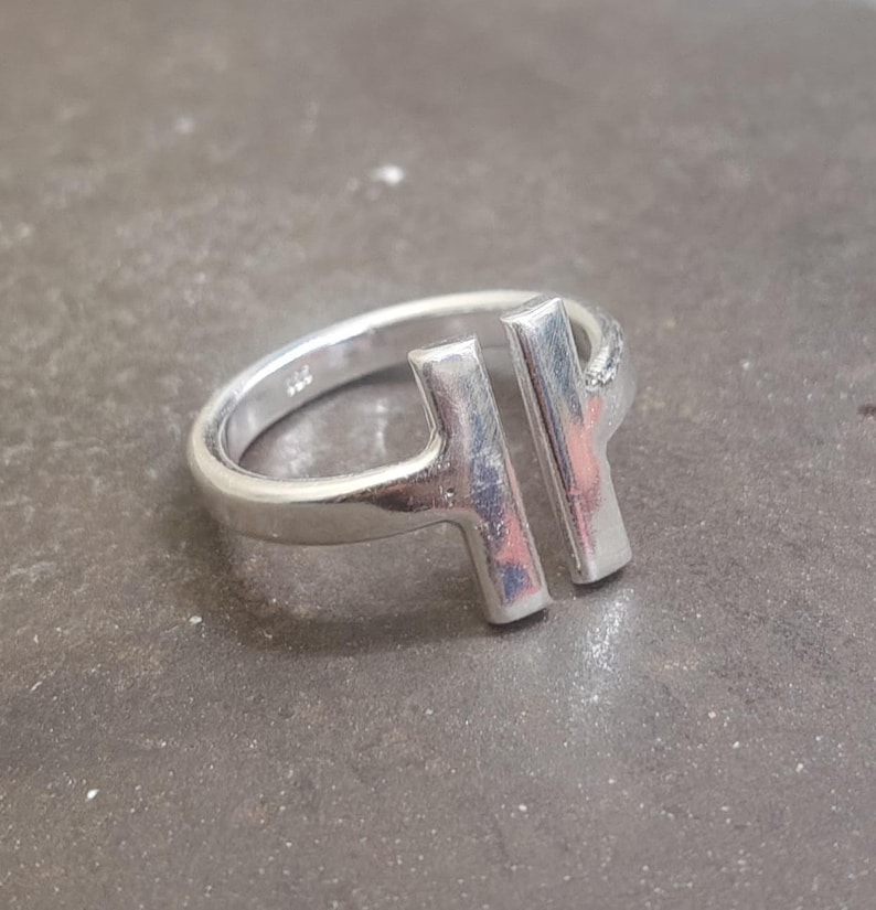 May include: A silver ring with a modern design. The ring features two vertical bars connected to the band, creating an open, geometric shape. The ring is made of a shiny, metallic material.