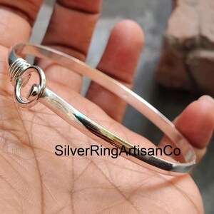 May include: A silver-colored bangle bracelet with a unique hook and loop closure. The bracelet is smooth and shiny, with a simple, elegant design. The text "SilverRingArtisanCo" is printed on the bracelet.