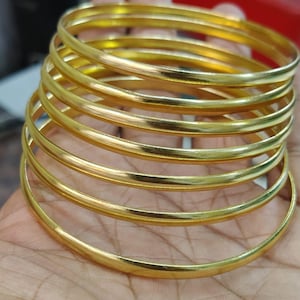 Puede incluir: Una pila de siete pulseras doradas, cada una con una superficie lisa y redondeada. Las pulseras están apiladas una encima de otra, creando un efecto de capas. Las joyas se exhiben sobre una superficie de color claro.