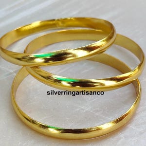 Peut inclure: Trois bracelets dorés s'entrelacent, reflétant la lumière. Les bandes lisses et arrondies sont empilées, créant un effet superposé. L'image comprend le texte "silverringartisanco".