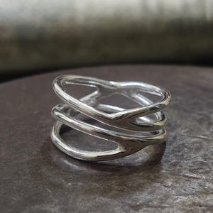 Op de afbeelding: Een zilveren ring met een gelaagd ontwerp. De ring heeft drie banden die elkaar kruisen en buigen, waardoor een open, geometrisch patroon ontstaat. Het metaal heeft een gepolijst, reflecterend oppervlak en de ring lijkt van sterling zilver te zijn.