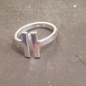 May include: A silver ring with a modern design. The ring features a band and two vertical bars, creating a unique T-shape. The ring is made of a shiny, metallic material, and is displayed on a textured surface.