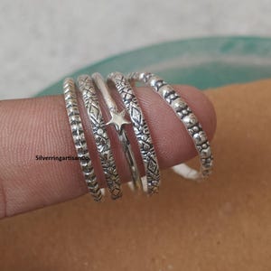 Può includere: Una collezione di anelli d'argento con vari design, tra cui motivi strutturati e un accento a forma di stella. Gli anelli sono mostrati su un dito, evidenziando i loro dettagli intricati. Gli anelli sono un accessorio delicato.