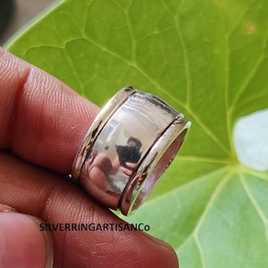 Può includere: Un anello color argento con una fascia rotante. L'anello ha una finitura lucida e un design strutturato sui bordi. L'anello è mostrato su una foglia verde.