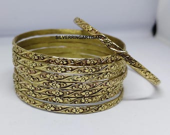 May include: A stack of gold-colored bangles with floral patterns. One bangle rests separately, displaying the intricate design. The bangles are stacked on a white surface. The text "SILVERRINGARTISANCO" is visible.