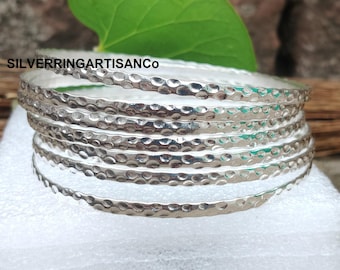 Peut inclure: Une pile de bracelets argentés avec une texture martelée. Les bracelets sont ronds et ont une surface brillante et réfléchissante. Le texte "SILVERRINGARTISANCO" est visible. Les bracelets sont empilés sur une surface blanche.
