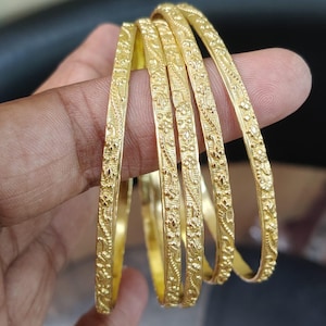 May include: A set of six gold bangles with intricate floral and leaf patterns. The bangles are a bright, polished gold color and appear to be made of metal. The design is consistent across all bangles, with detailed carvings.