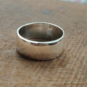Può includere: Un anello largo e lucido in argento con una fascia liscia e arrotondata. L'anello riflette la luce, creando riflessi e ombre. Il gioiello è appoggiato su una superficie di legno, esaltando la sua lucentezza metallica.