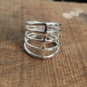 Puede incluir: Un anillo plateado con un diseño en forma de jaula. El anillo presenta múltiples bandas horizontales conectadas por barras verticales, creando un patrón geométrico abierto. El anillo está hecho de un metal brillante y reflectante.