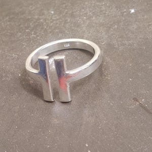 May include: A silver ring with a modern design. The ring features a band and a vertical bar design. The ring is made of a shiny, metallic material. The ring is on a gray surface.