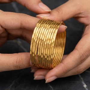 Könnte beinhalten: Ein Set aus zehn goldfarbenen Armreifen mit einer gehämmerten, strukturierten Oberfläche. Jeder Armreif hat ein dünnes, abgerundetes Profil mit einer reflektierenden metallischen Oberfläche, die im Verbund einen gestapelten Look erzeugt.