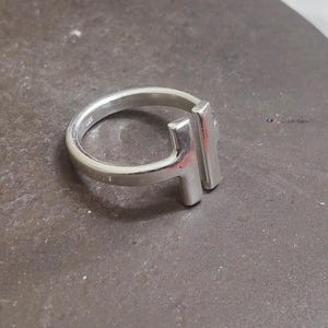 May include: A silver ring with a modern design. The ring features a rounded band and a rectangular bar element. The ring is made of a shiny, metallic material and is set against a neutral background.