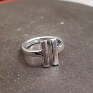 May include: A silver ring with a modern design. The ring features two vertical bars connected to the band, creating a minimalist and geometric aesthetic. The ring is made of a shiny, reflective metal.
