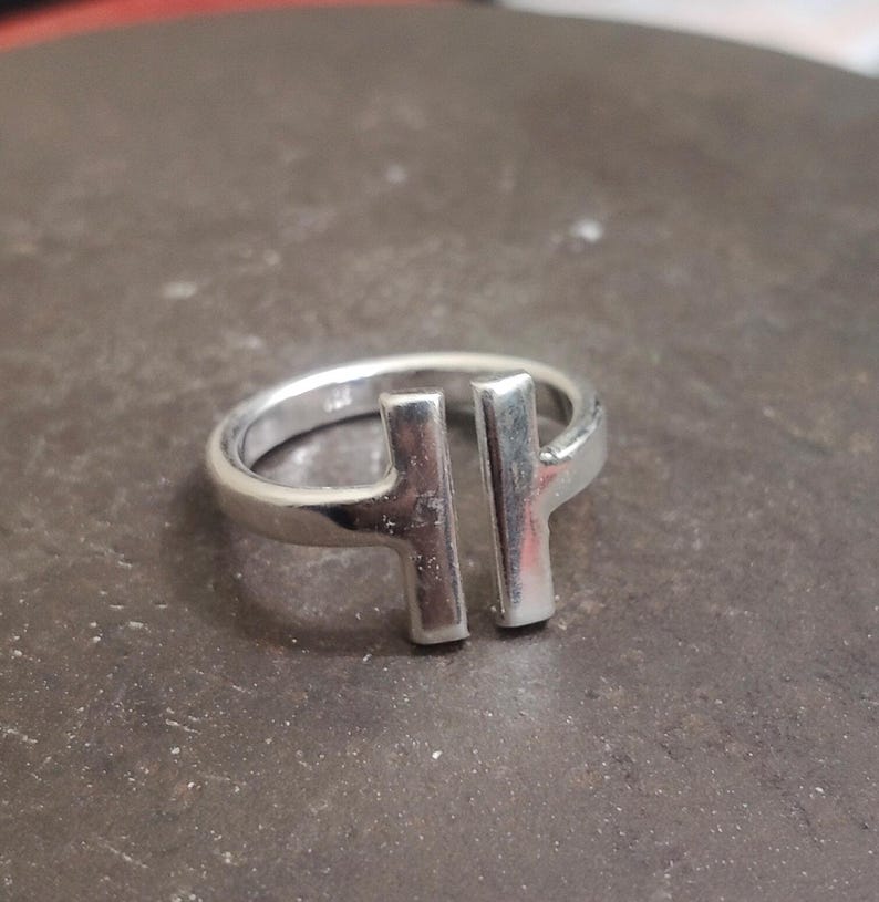May include: A silver ring with a modern design. The ring features two vertical bars connected to the band, creating a minimalist and geometric aesthetic. The ring is made of a shiny, reflective metal.