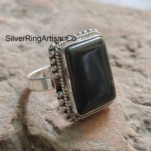 May include: A silver ring with a large, rectangular black stone. The ring has a detailed, textured border and a simple band. The text "SilverRingArtisanCo" is visible in the image.