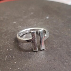 May include: A silver ring with a modern design. The ring features two vertical rectangular bars connected by a band. The ring is made of a shiny, reflective metal.