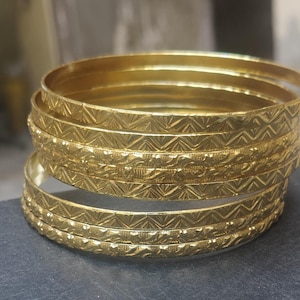 Op de afbeelding: Een stapel goudkleurige armbanden. Elke armband heeft een zich herhalend geometrisch patroon dat in het oppervlak is geëtst. De armbanden zijn op elkaar gestapeld, waarbij de bovenste armband enigszins onscherp is. Ze liggen op een donkere ondergrond.