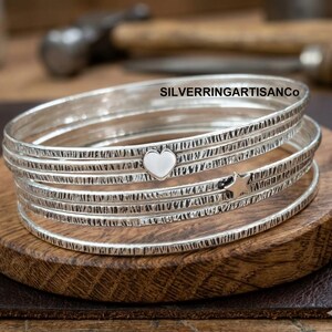 May include: A stack of five silver bangles with a textured, hammered finish. One bangle features a small, smooth silver heart charm, and another displays a small, smooth silver star charm. The text SILVERRINGARTISANCo is visible.