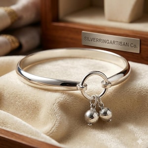 Puede incluir: Una pulsera rígida de plata pulida con un elemento de anillo central. Dos pequeños dijes esféricos texturizados cuelgan del anillo. Una placa de metal en el fondo muestra el texto SILVERRINGARTISAN Co. en una fuente clara.