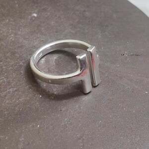 May include: A silver ring with a modern design. The ring features a rounded band and a rectangular bar element. The ring is made of a shiny, metallic material and is set against a neutral background.