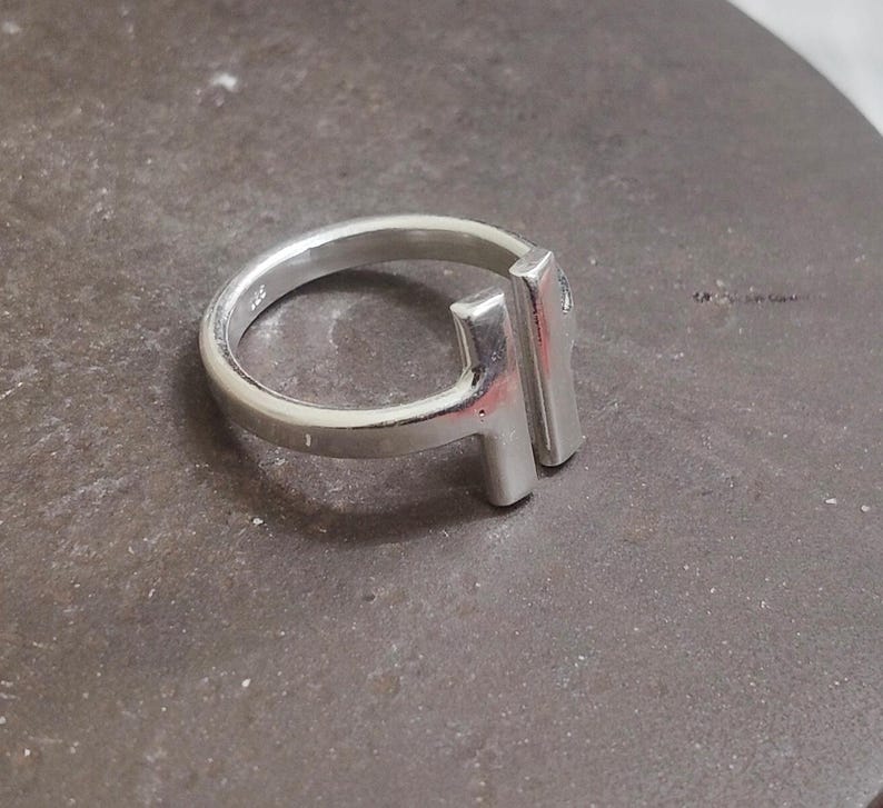 May include: A silver ring with a modern design. The ring features a rounded band and a rectangular bar element. The ring is made of a shiny, metallic material and is set against a neutral background.