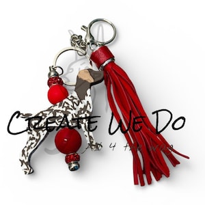 May include: A keychain featuring a dog-shaped charm with a black and white spotted pattern, a red tassel, and red beaded accents. The keychain includes the text "Create We Do".