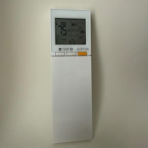 May include: A white air conditioning remote control with a digital display showing 75°F and the time 9:12 AM. The remote has buttons for temperature and on/off functions.
