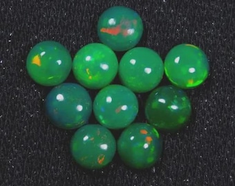 May include: Ten polished, round green opals are displayed. The opals have a glossy finish with internal colour variations of yellow, orange, and red. They are arranged closely together against a dark, textured background.
