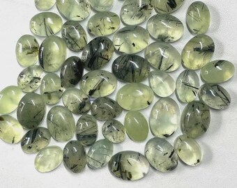 May include: A collection of oval-shaped prehnite gemstones. The stones vary in size and have a translucent, pale green color with dark inclusions. The gemstones are arranged on a white surface, showcasing their natural beauty and unique patterns.