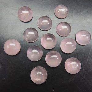 Könnte beinhalten: Eine Sammlung von elf polierten, runden Rosenquarzsteinen. Jeder Stein hat einen zarten rosa Farbton und eine glatte, reflektierende Oberfläche. Die Steine sind auf einer dunklen Oberfläche angeordnet, was ihre natürliche Farbe und Form hervorhebt.