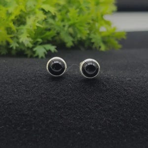 Puede incluir: Un par de pendientes de botón con piedras negras redondas, lisas y pulidas, engastadas en un marco de metal de tono plateado. Los pendientes tienen un diseño circular con un acabado brillante y un color oscuro sólido que resalta el borde.