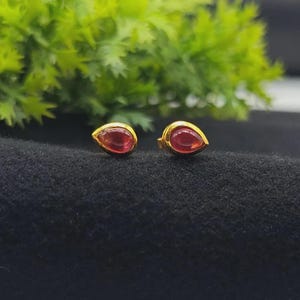 Puede incluir: Un par de pendientes de botón con gemas rojas en forma de lágrima engastadas en metal dorado. Cada pendiente tiene un acabado liso y pulido con un extremo puntiagudo, mostrando un tono carmesí vibrante que contrasta con el engaste metálico.