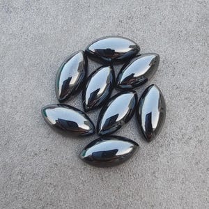 May include: A collection of eight polished, oval-shaped hematite stones. The stones have a dark, metallic gray color and a smooth, reflective surface. They are arranged on a gray, textured surface.