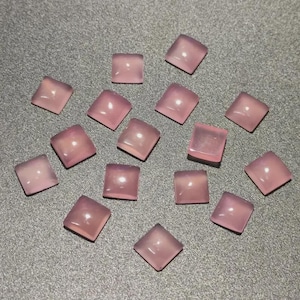 Puede incluir: Un conjunto de gemas cuadradas de color rosa claro con esquinas redondeadas. Las gemas están dispersas sobre una superficie gris, cada una reflejando la luz. Las piedras parecen pulidas y translúcidas, mostrando un color pastel suave.