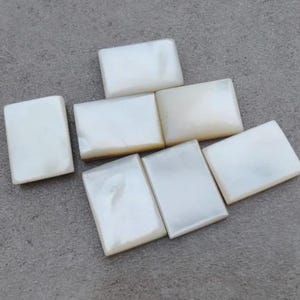 May include: Seven rectangular, off-white mother-of-pearl tiles are arranged on a gray surface. The tiles have a smooth, glossy finish and a slightly iridescent sheen. The tiles are likely for jewelry making or craft projects.