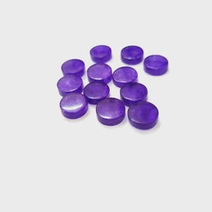 May include: A collection of small, round, polished purple gemstones. The gemstones are scattered across a white surface, each with a flat top and bottom. The color is a deep, rich purple, and the stones appear smooth and slightly translucent.
