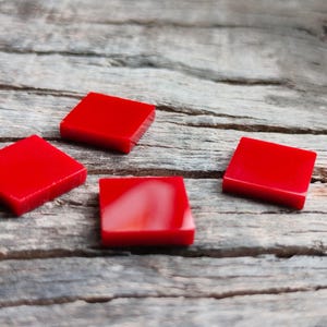 Op de afbeelding: Vier kleine, vierkante, felrode tegels zijn gerangschikt op een verweerd, grijs houten oppervlak. Eén tegel heeft een witte werveling. De tegels zijn gelijkmatig verdeeld en de houtnerf zorgt voor een textuurachtergrond.